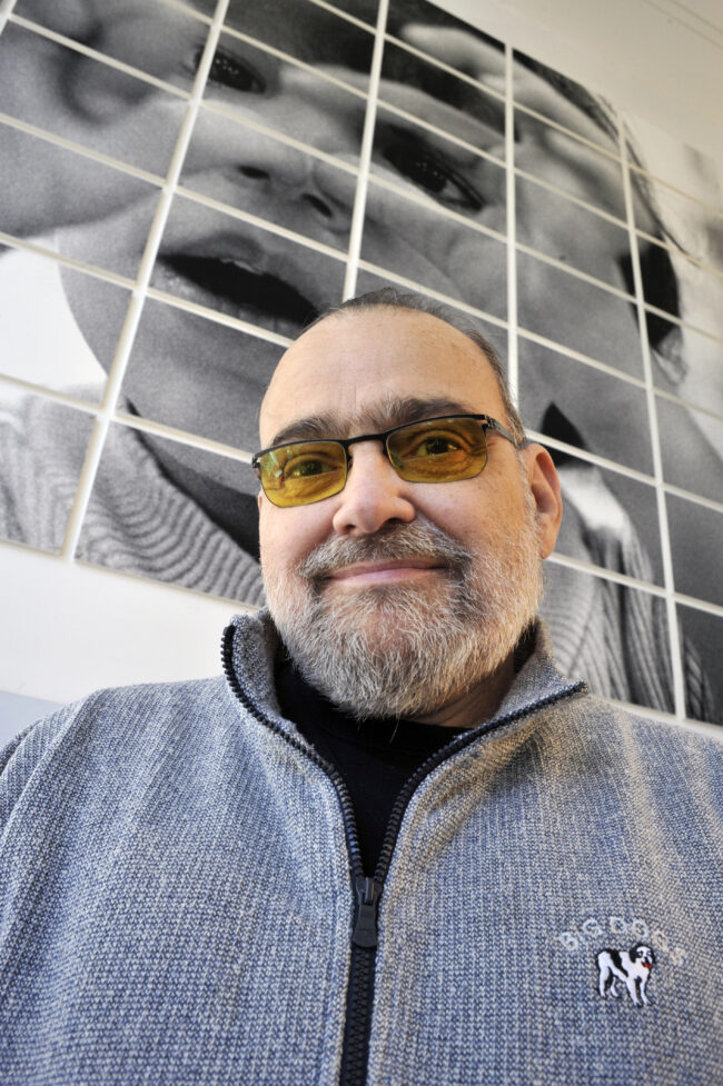 Handsome, middle aged man with a full gray beard and yellow-tinted glasses smiles at the camera. He wears a gray zip-up sweater with a 